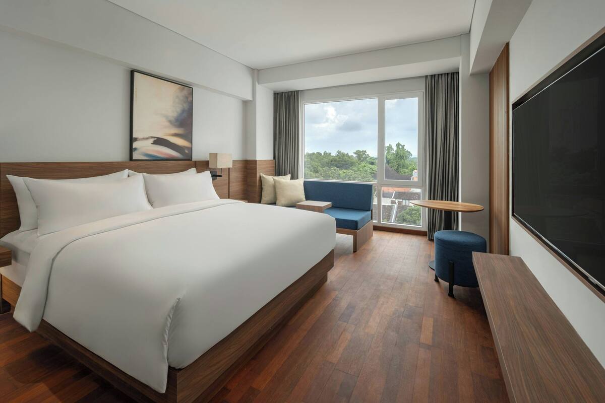 A modern hotel room with a large bed, a seating area with a small table, a large window with curtains, and a flat-screen TV.