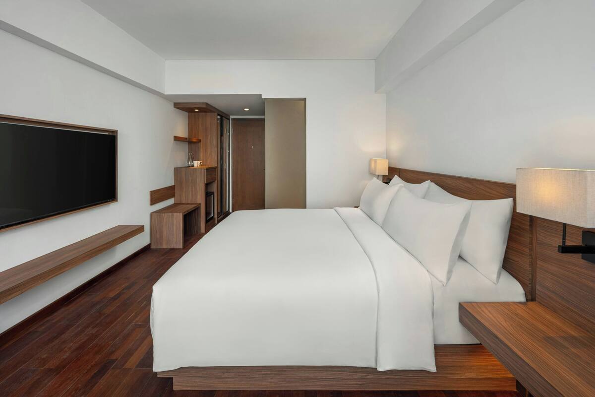A modern hotel room with a large bed, wooden headboard, bedside tables, a flat-screen TV, and built-in shelves.