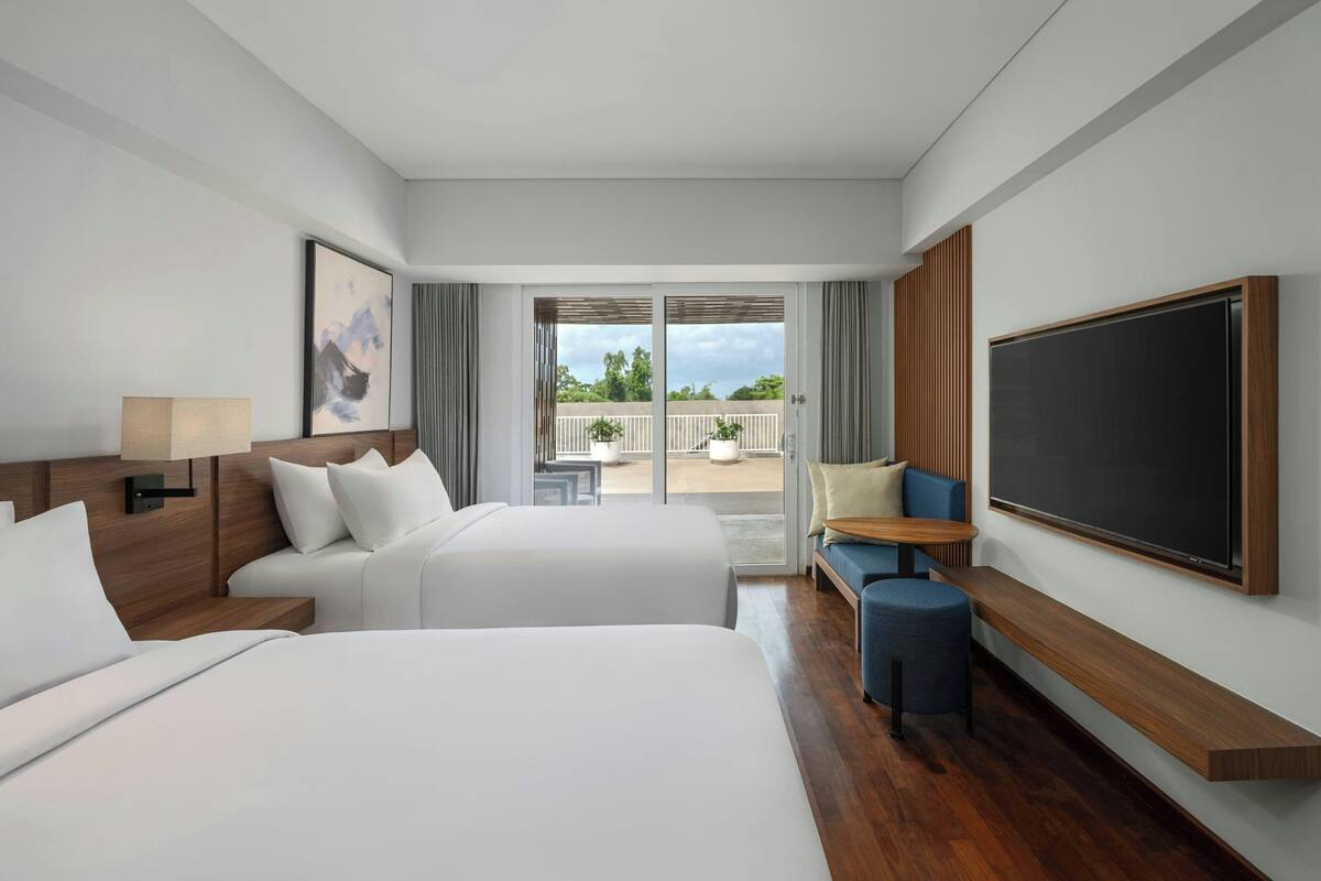 A hotel room with two beds, a large flat-screen TV, a balcony with a view, and a seating area with a table.