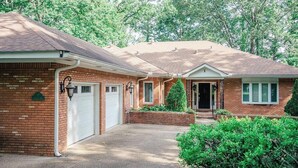 A two-story brick house with a garage, a front porch, and a small front yard with a tree and bushes.