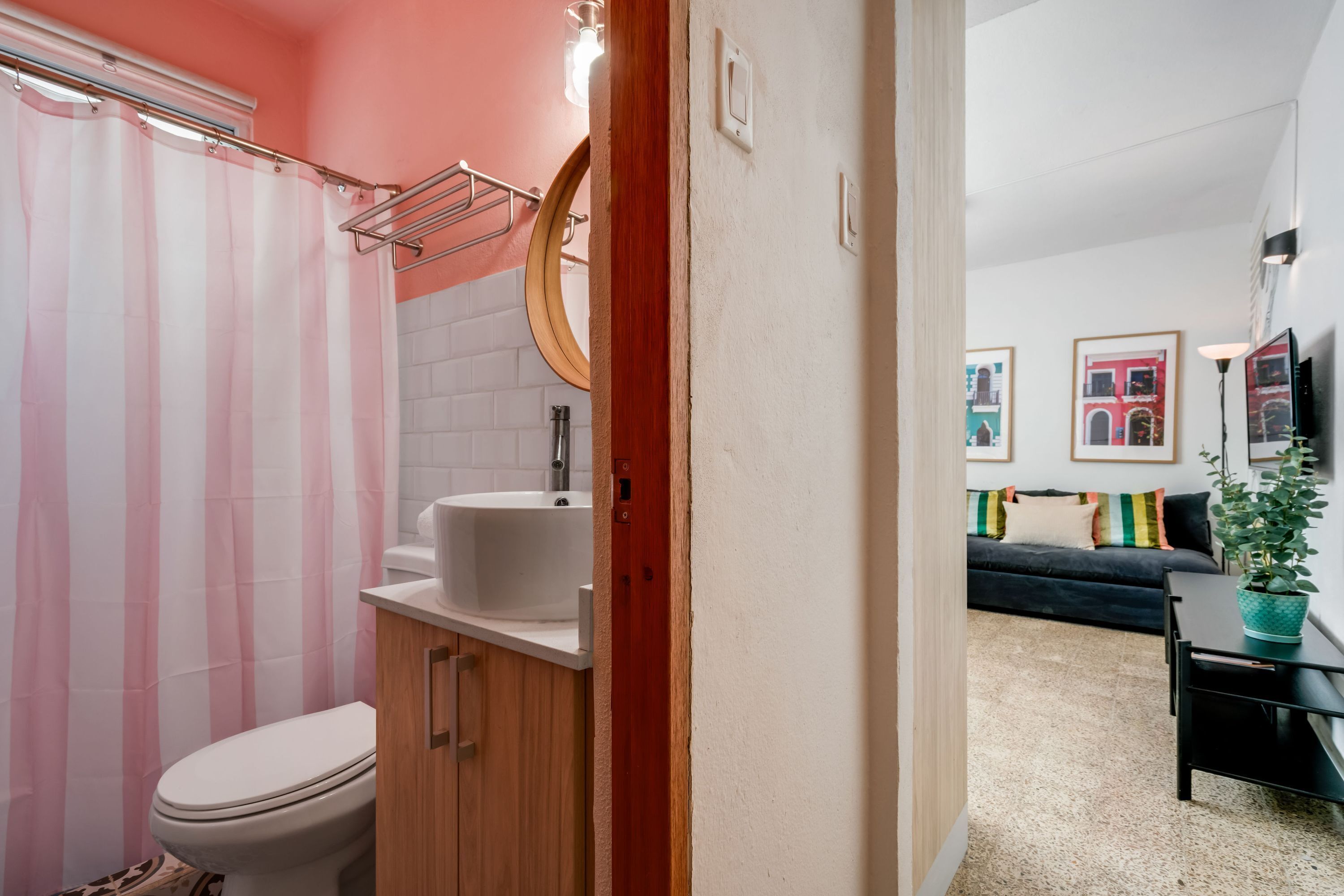 A bathroom with a shower curtain, sink, and toilet. There is a wooden cabinet and a mirror above the sink. The room has a beige floor and a dark-colored sofa in the background.