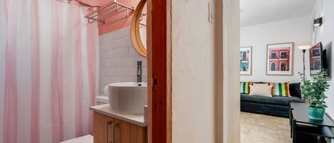 A bathroom with a shower curtain, sink, and toilet. There is a wooden cabinet and a mirror above the sink. The room has a beige floor and a dark-colored sofa in the background.