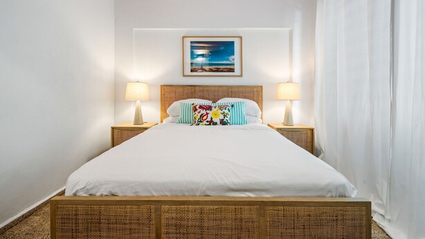 A neatly made bed with white linens, two bedside lamps, a framed picture above the headboard, and a white curtain on the right. - Oasis Condado Pool Parking Beach Steps Away (San Juan)