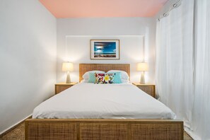 Un lit bien fait, avec du linge de lit blanc, deux lampes de chevet, un tableau encadré au-dessus de la tête de lit et un rideau blanc sur le côté droit.