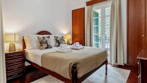 Una cama bien hecha con una colcha color beige, almohadas y toallas dobladas. Hay una mesita de noche de madera con una lámpara, una ventana con cortinas y vistas a la vegetación.