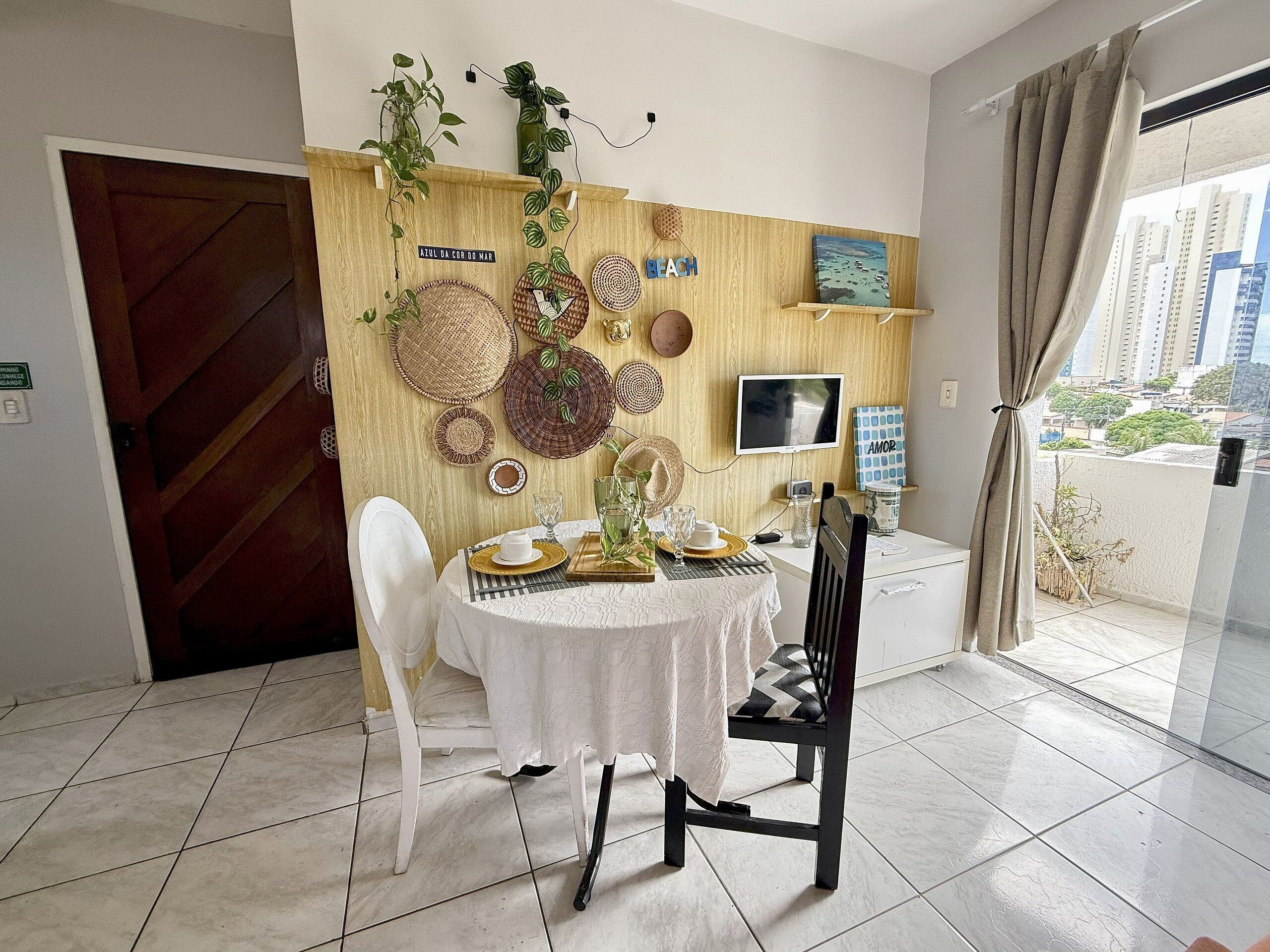 A dining area with a table set for two, a TV on the wall, and a view of the city through the window.
