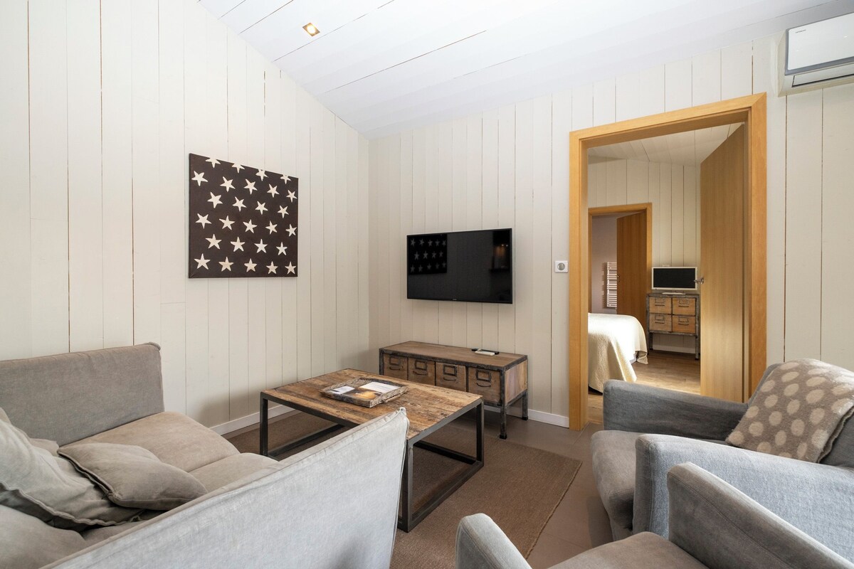 Een moderne woonkamer met een bank, fauteuil, salontafel en een flatscreen televisie aan de muur. Er is een houten kast met lades en een klein tafeltje met een dienblad.