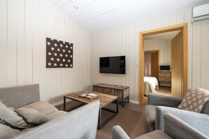 A modern living room with a sofa, armchair, coffee table, and a flat-screen TV mounted on the wall. There is a wooden cabinet with drawers and a small table with a tray. - Mv Lodge (La Couarde-sur-Mer)