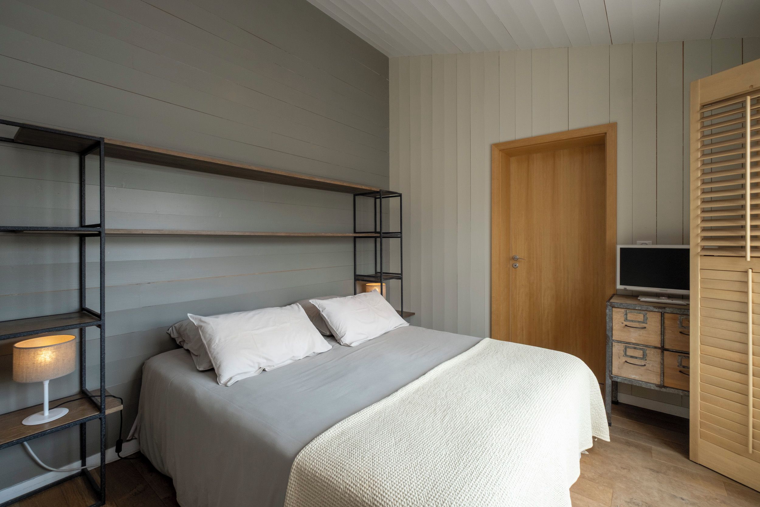 A bedroom with a bed, a nightstand with a lamp, a wooden wardrobe, and a TV stand.