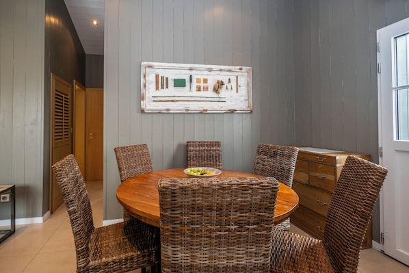 A dining area with a round wooden table surrounded by wicker chairs, a rustic wall art piece above, and a wooden cabinet to the right. - Mv Lodge (La Couarde-sur-Mer)