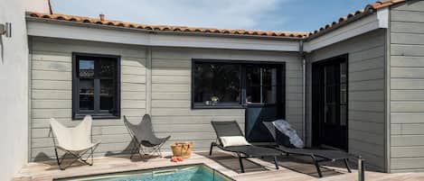 A poolside area with a wooden deck, two lounge chairs, a small table with a basket, and a window.