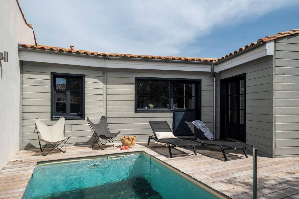 Un espace aménagé au bord de la piscine, avec une terrasse en bois, deux chaises longues, une petite table sur laquelle se trouve un panier, et une fenêtre.