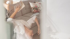 Eine Person liegt entspannt auf einem Sofa, mit einem Buch, einem Blumenkissen und einer Pflanze im Hintergrund.