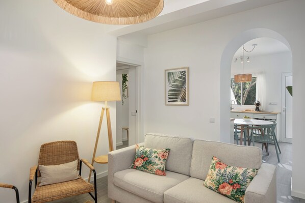 A modern living room with a sofa, armchair, and floor lamp. There is a dining area visible through an archway.