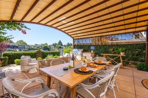 Eine Terrasse im Freien mit Holztisch, Korbstühlen, einem Grill und Blick auf eine Wohngegend.