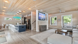 A spacious living area with a wooden ceiling, a large TV mounted on a wall, a kitchen area, and a seating area with a sofa and chairs.