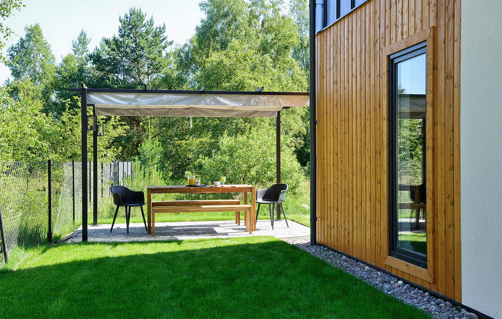 A patio with a wooden table and chairs, a pergola, and a view of a forested area.