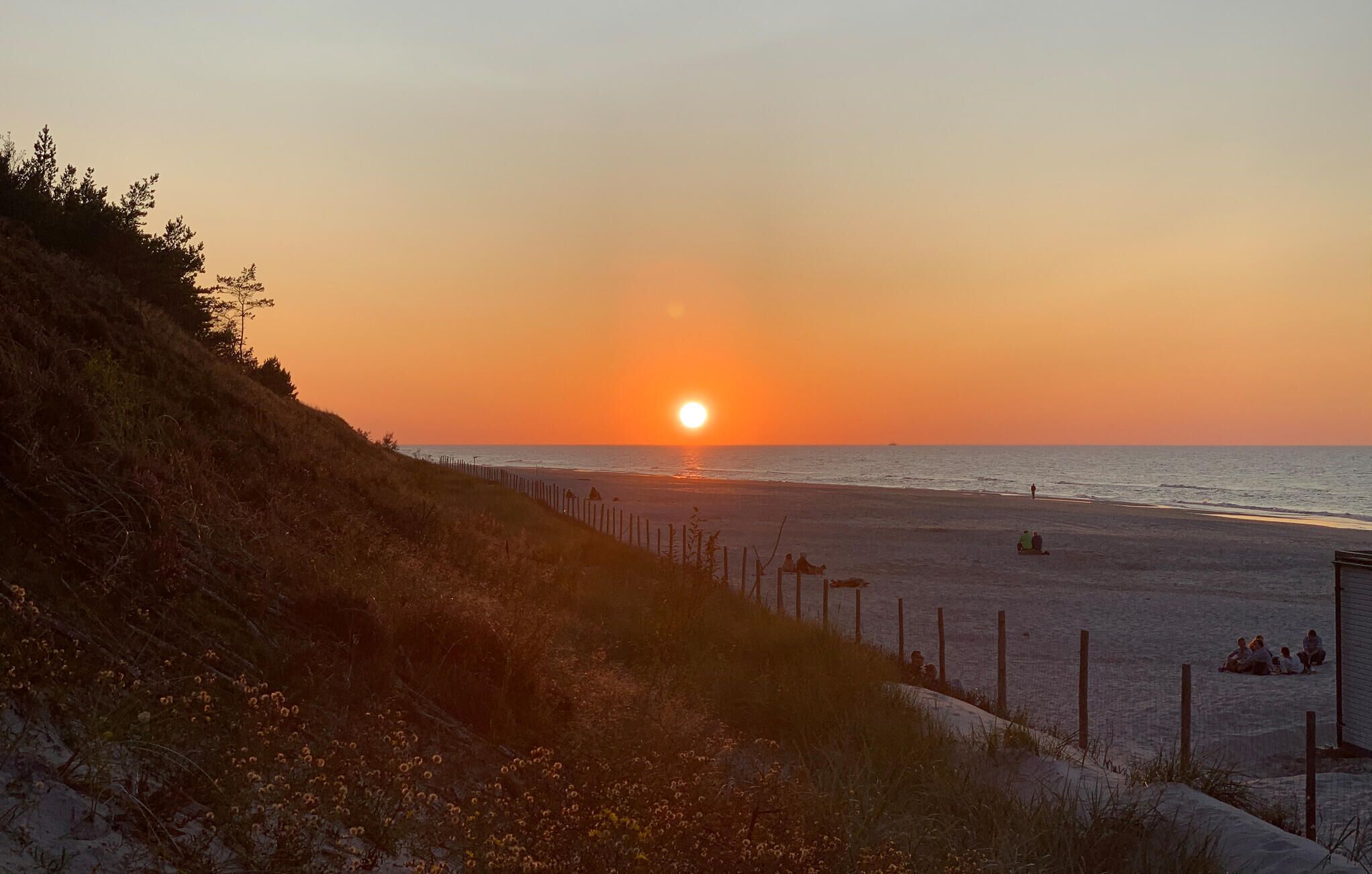 Un atardecer sobre una playa con una valla y gente sentada en la arena.