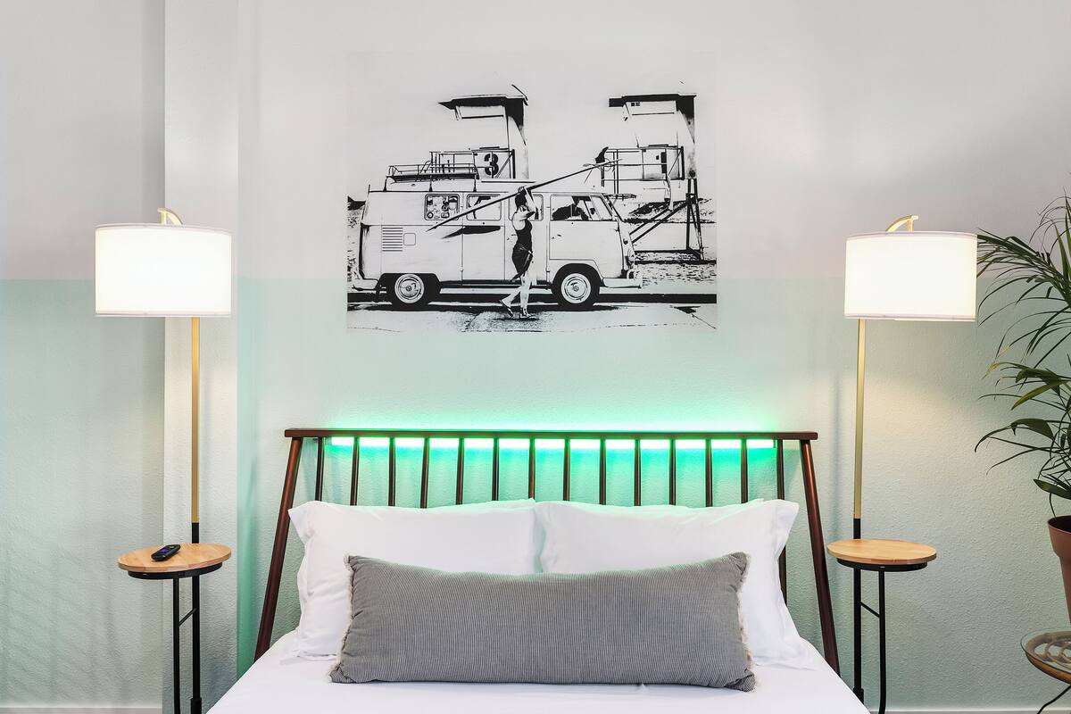 Camera d'albergo con un letto, due comodini, una lampada a parete, una testiera illuminata di verde e un'opera d'arte in bianco e nero raffigurante un carro attrezzi.