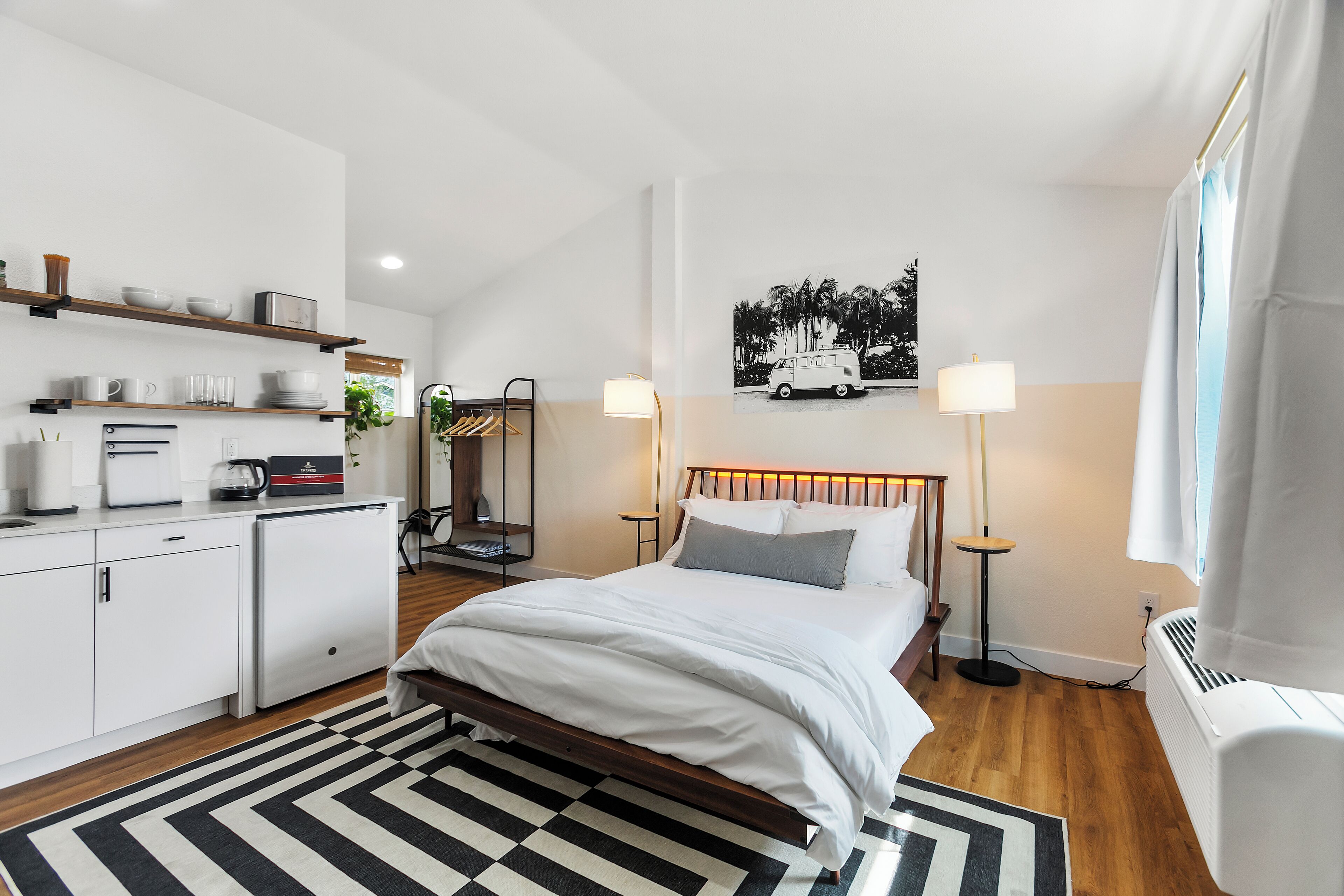 A modern hotel room with a bed, bedside lamps, a coffee machine, and a kitchen area with shelves.