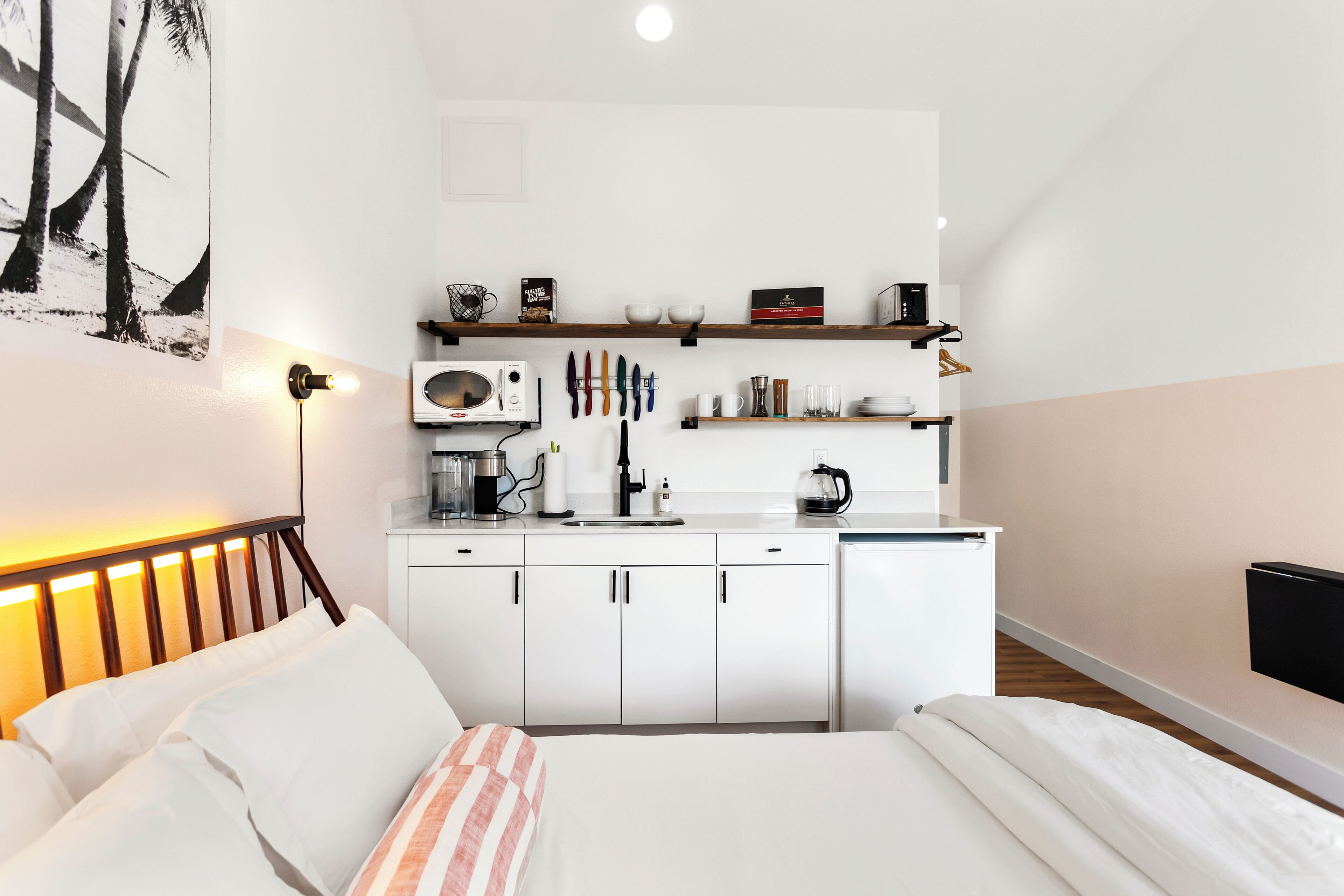 A modern hotel room with a bed, bedside table, and a kitchenette area with a microwave, coffee maker, and utensils.