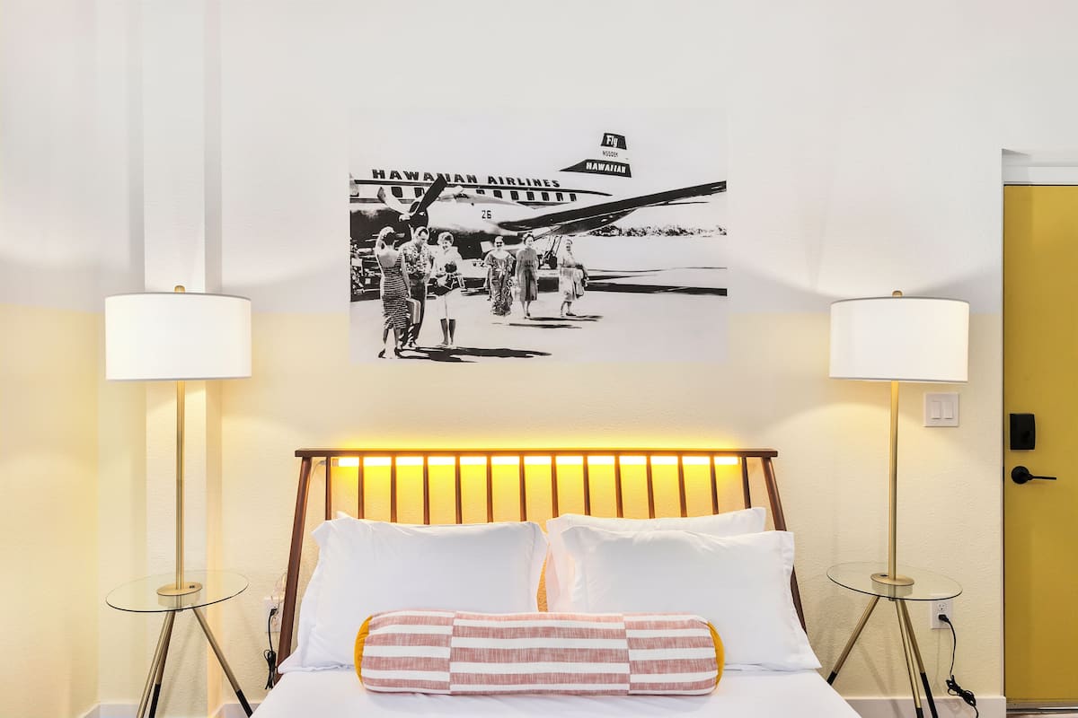 Una camera d'albergo con un letto, due lampade sui comodini, un quadro appeso al muro raffigurante Hawaiian Airlines e una porta gialla.