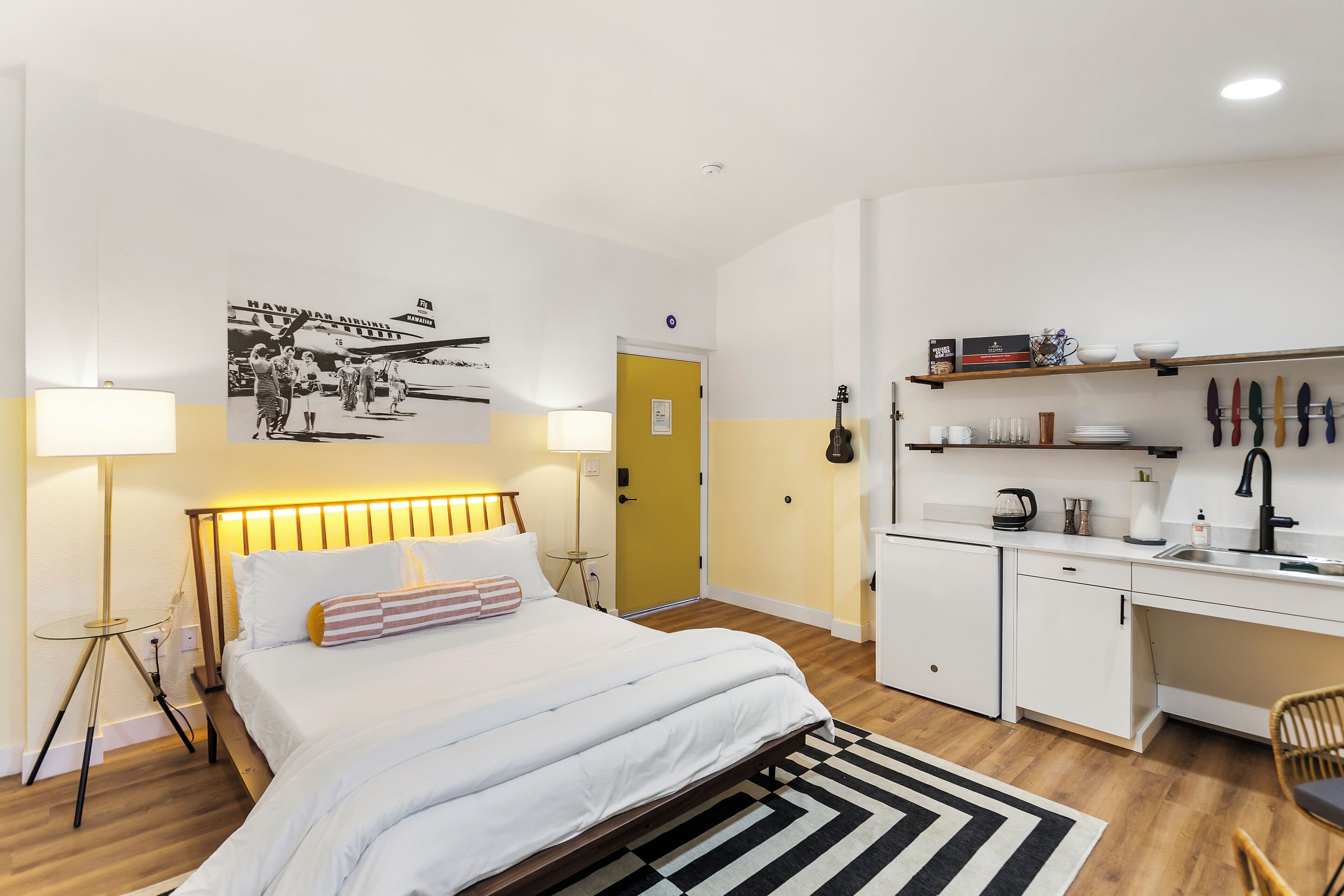 Une chambre d’hôtel moderne équipée d’un lit, d’une table de chevet, d’une lampe et d’un coin cuisine comprenant un évier, des placards et des ustensiles.
