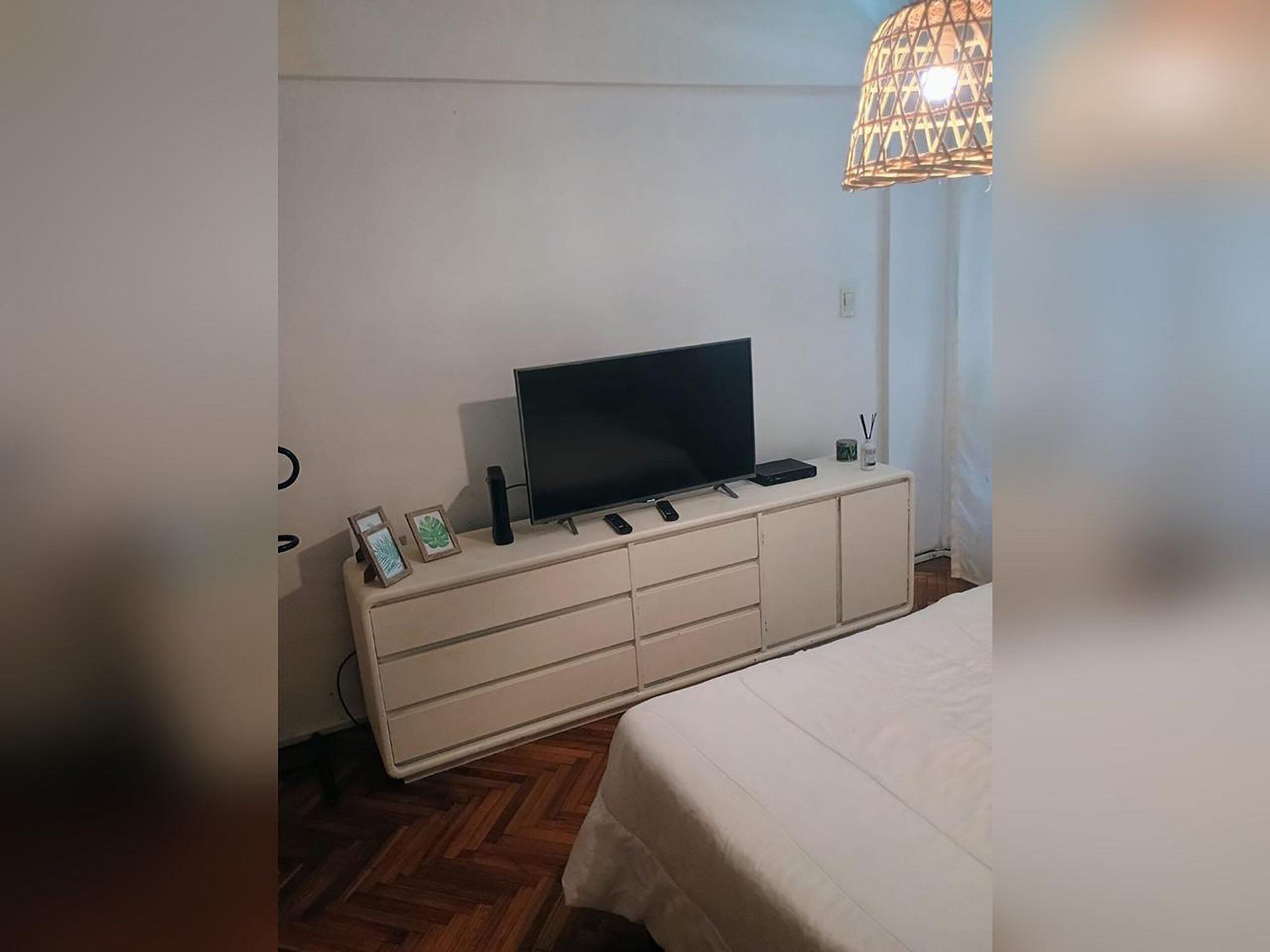 Een slaapkamer met een bed, een televisie op een witte dressoir, een lamp en een klein tafeltje met een plant.