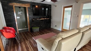 A modern living room with a wooden floor, a beige sofa, red chairs, a flat-screen TV mounted on a dark wall, and a ceiling fan.