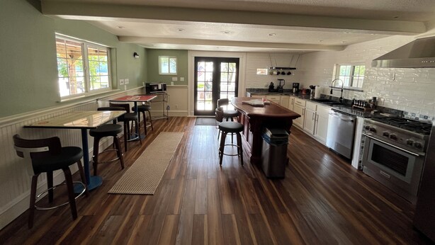 A spacious kitchen with wooden flooring, a central island, bar stools, a dining table, and a large stove.