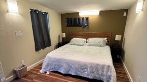 A small, neatly arranged bedroom with a wooden floor, a bed with a white quilt, two bedside tables with lamps, and a window with curtains.
