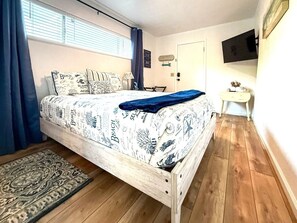 Un dormitorio con una cama de madera, una colcha estampada, un plaid azul, una mesita blanca pequeña y un televisor en la pared.