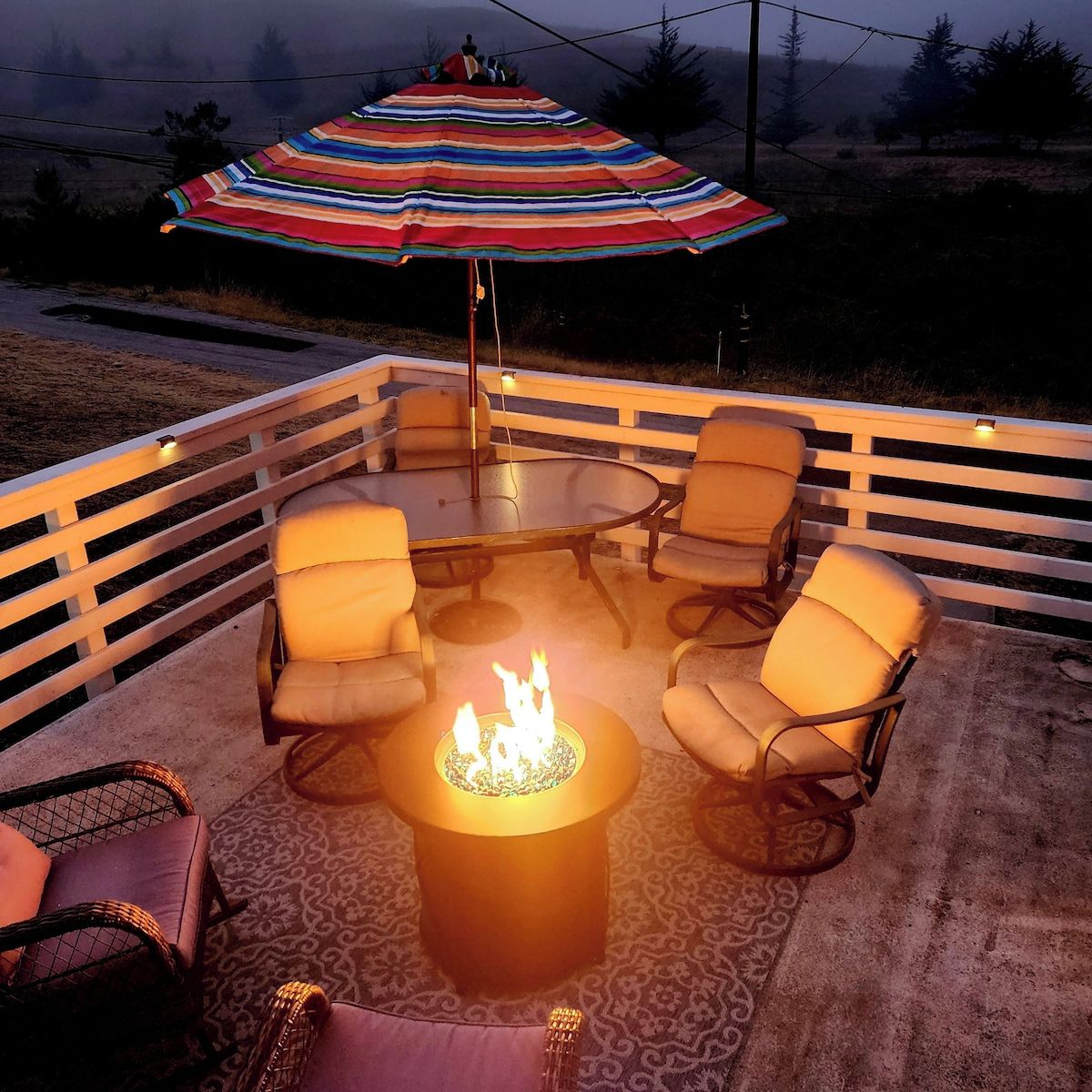 Een terras buiten met een ronde tafel en stoelen, een vuurkorf en een strepenpatroon parasol.