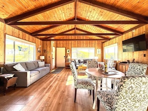 A spacious wooden interior with a sofa, patterned chairs, a dining table, and a television.