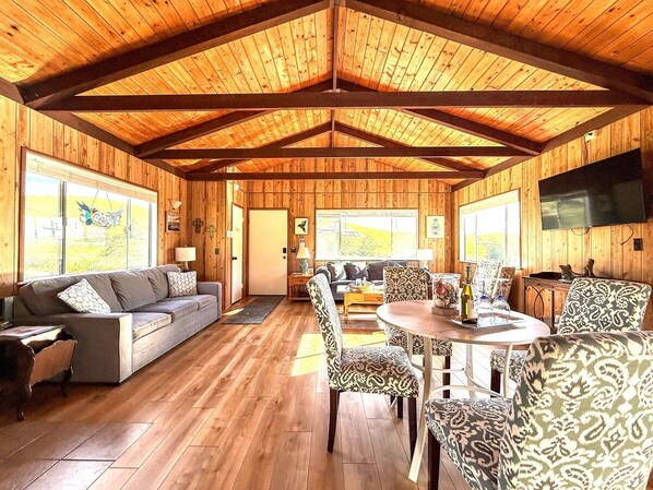 A spacious wooden interior with a sofa, patterned chairs, a dining table, and a television.