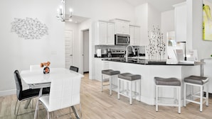 A modern kitchen with white cabinetry, a central island with bar stools, and a dining area with a white table and chairs. - Newley Built/Heated Pool/Golf Course Views/Sleeps 9 (Rotonda West)