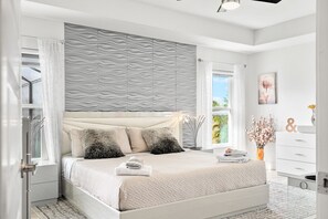 Une chambre moderne avec un grand lit, une tête de lit intégrée, une table de chevet, une commode, une fenêtre avec des rideaux, un vase avec des fleurs et une œuvre d’art accrochée au mur.