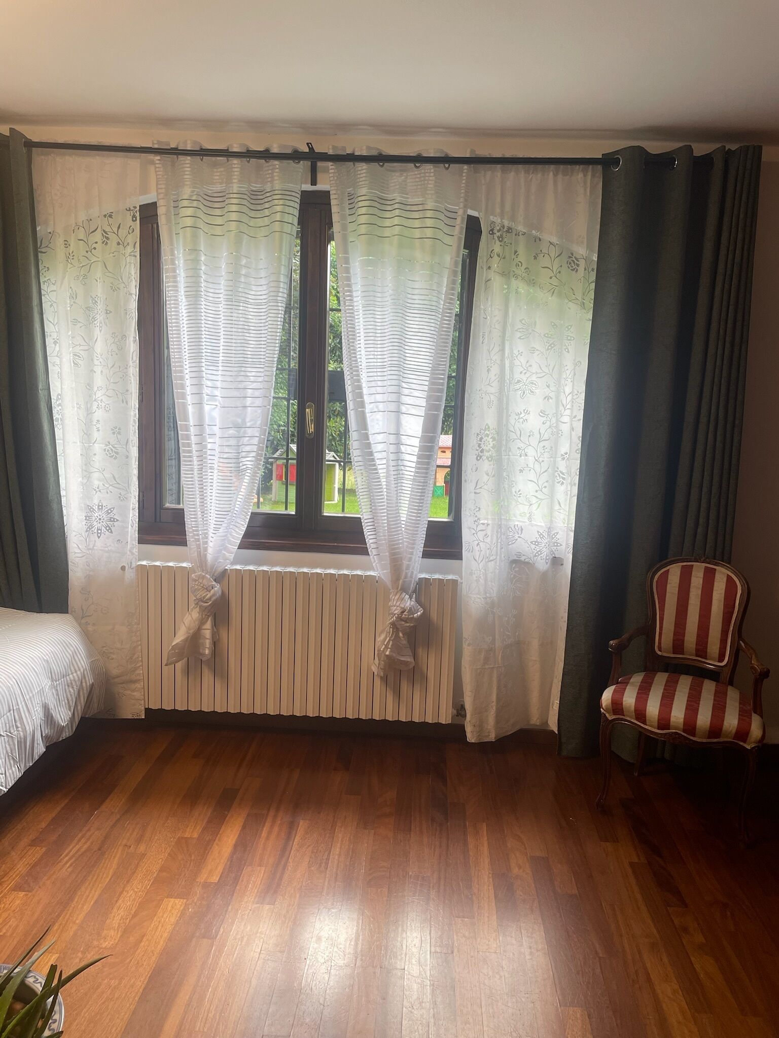 Quarto com piso de madeira, radiador, cadeira e janela com cortinas.
