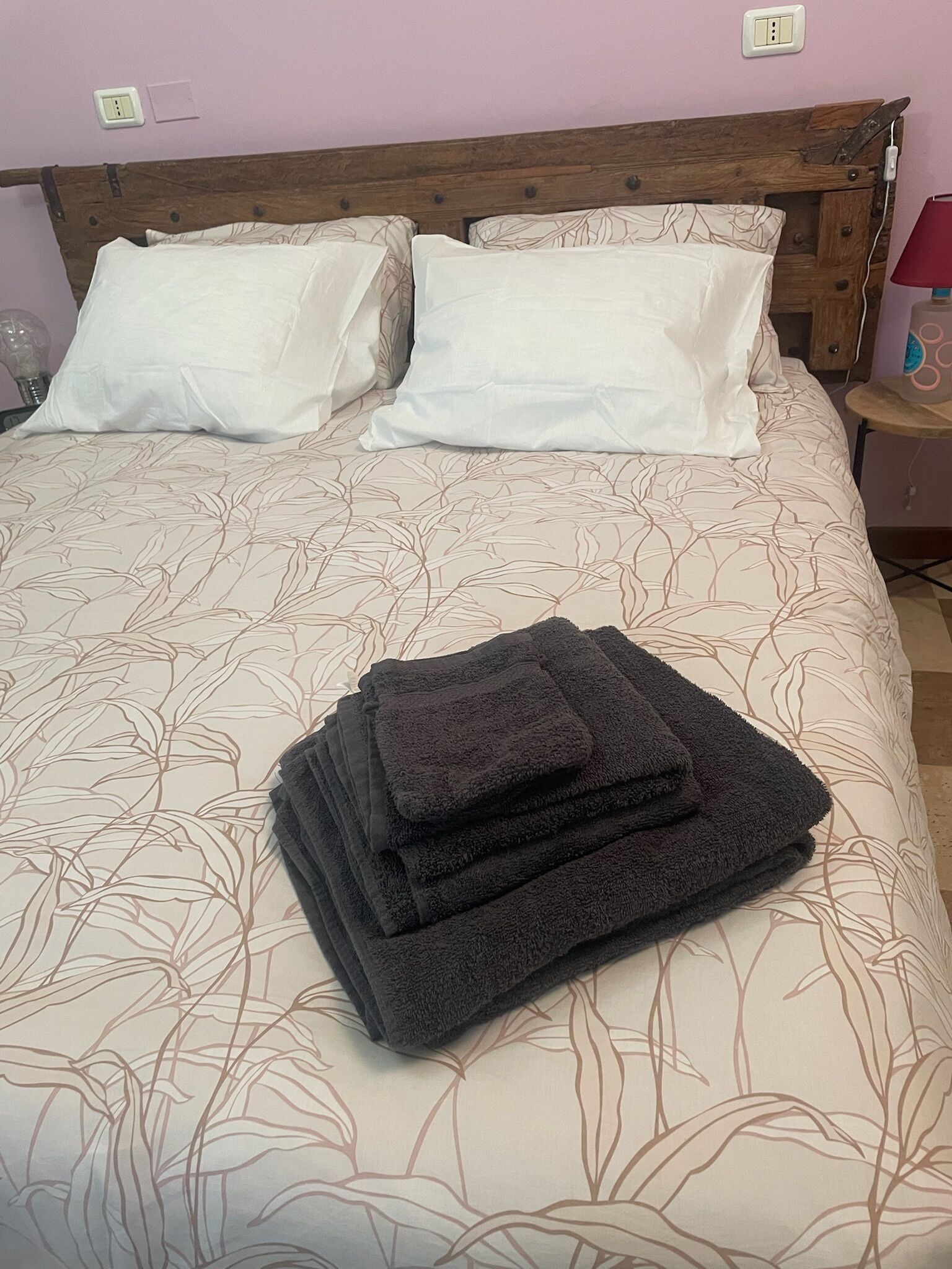 Ein ordentlich bezogenes Bett mit einem blumigem Überwurf, zwei weißen Kissen und einem Stapel gefalteter dunkler Handtücher.