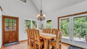 Une salle à manger avec une table et des chaises en bois, un lustre et de grandes fenêtres donnant sur un paysage verdoyant.