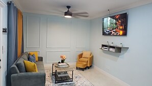 A living room with a grey sofa, a yellow armchair, a coffee table, and a TV mounted on the wall displaying a cityscape. - Charming Modern 1-bedroom apartment with WiFi, AC in Beautiful Nassau (Nassau)