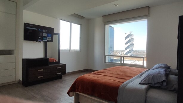 A bedroom with a bed, a dresser, a TV mounted on the wall, and a window with a view of a building. - Apartment in Sonata, Puebla (Heroica Puebla de Zaragoza)