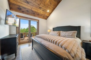 Une chambre avec un plafond en bois, un grand lit recouvert d’une couverture moelleuse, une table de chevet, un téléviseur fixé au mur et une porte-fenêtre coulissante donnant sur une terrasse extérieure.