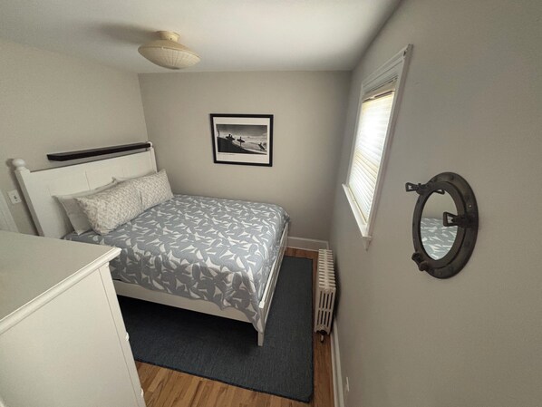A bedroom with a bed, a bedside table, a radiator, a window with blinds, and a framed picture on the wall. - Cozy and charming, this cottage offers a retreat surrounded by local beaches. (Portsmouth)