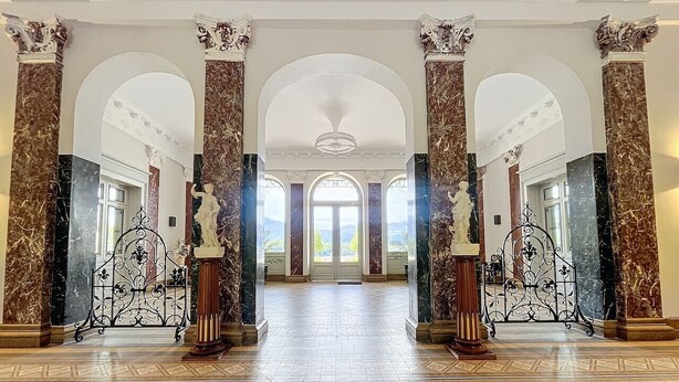 A grand hallway with marble columns, ornate metal gates, and a patterned floor leading to a large arched doorway.