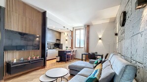 A modern living room with a sofa, ottoman, and a round table. A kitchen is visible in the background with a dining area.