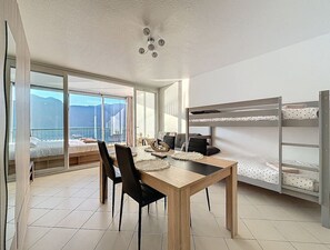 Une chambre moderne avec une table à manger en bois, des chaises noires, un lit superposé avec une literie blanche et de grandes fenêtres offrant une vue sur la montagne.