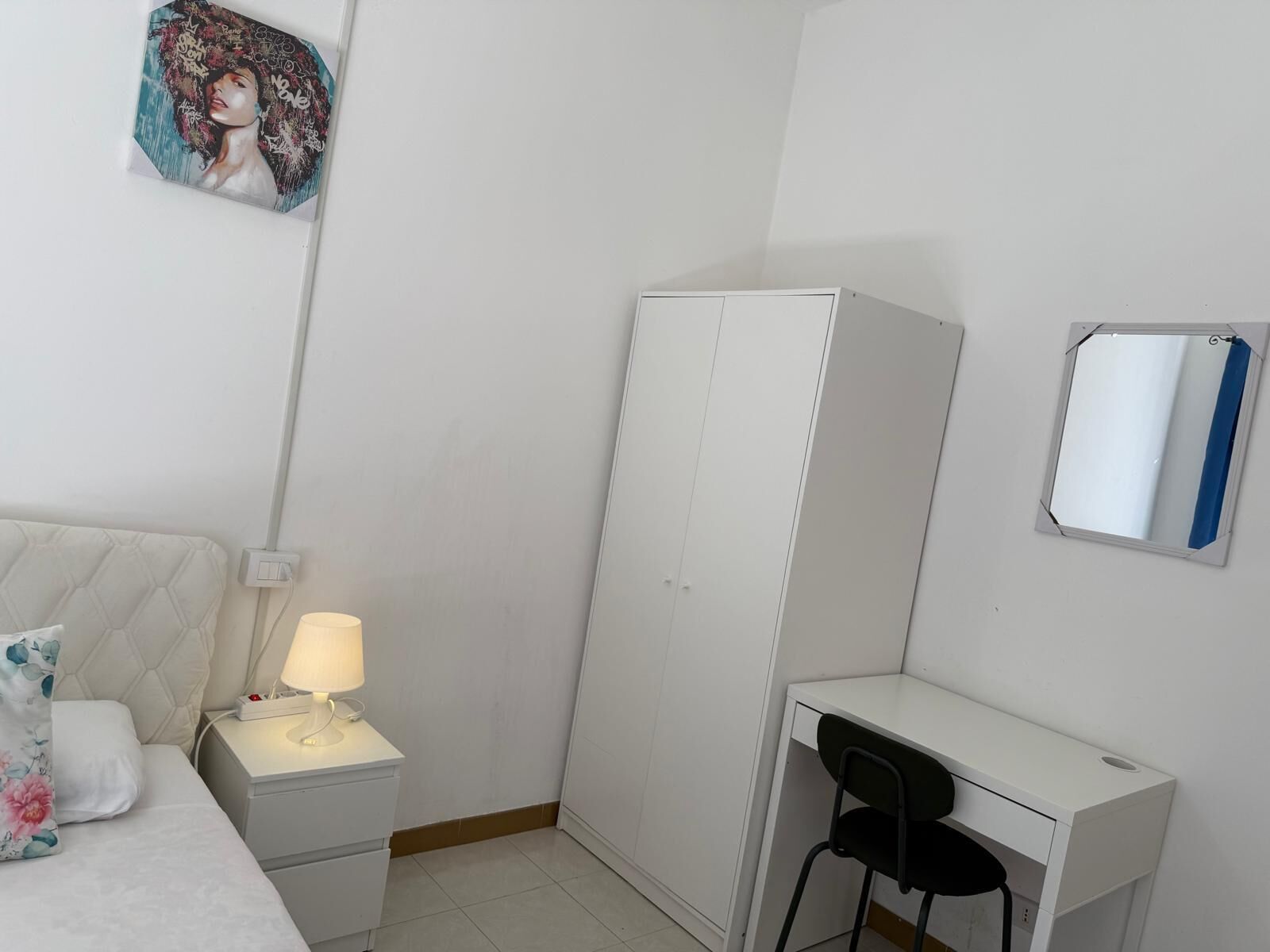 A bedroom with a bed, nightstand, desk, chair, wardrobe, and a wall-mounted mirror.