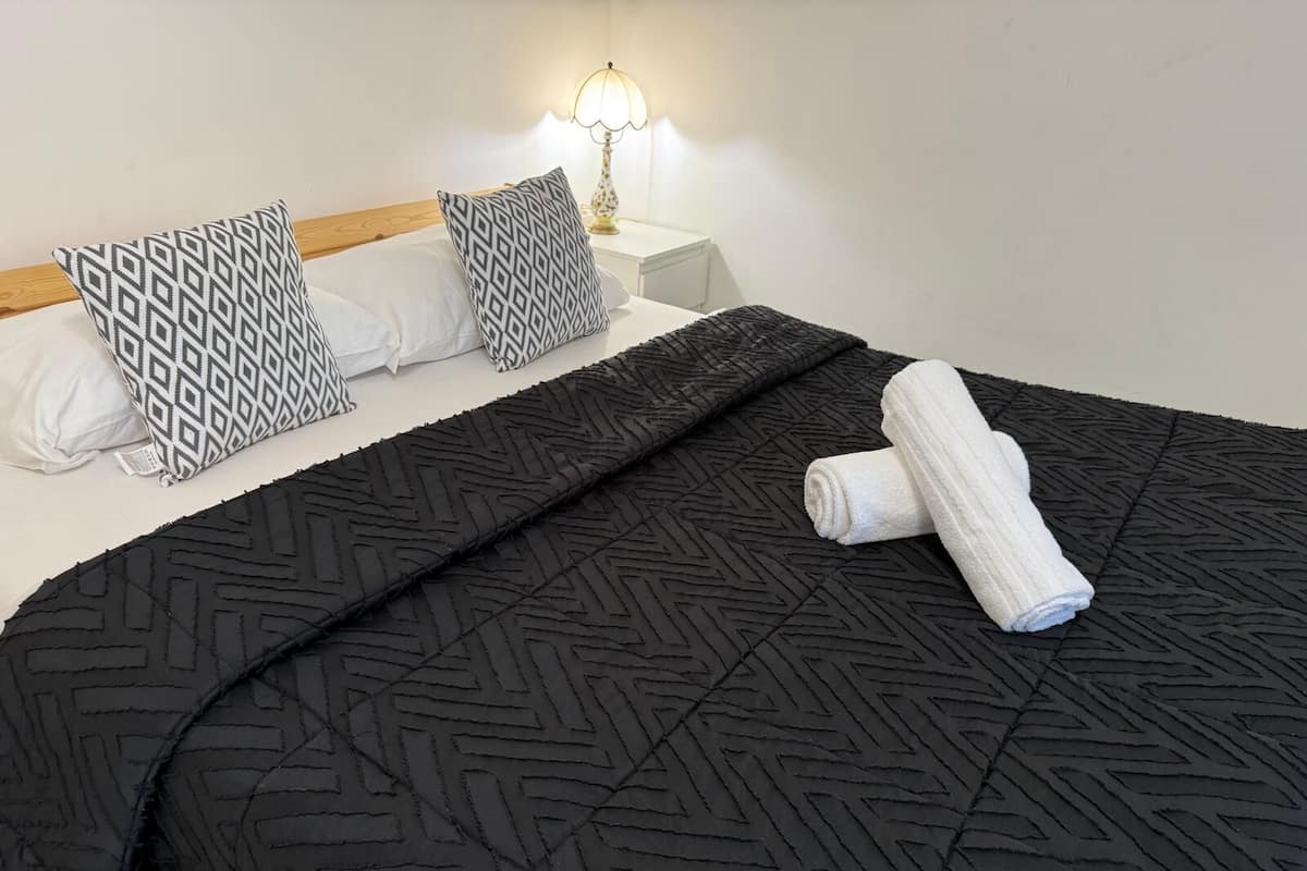 A neatly made bed with a black geometric-patterned bedspread, two patterned pillows, a rolled white towel, and a bedside lamp on a white nightstand.
