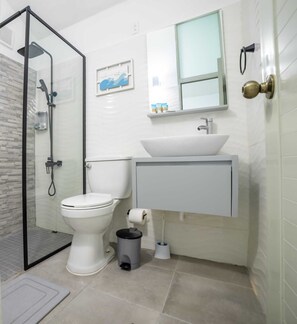 Un baño moderno con cabina de ducha de vidrio, inodoro blanco, lavamanos con espejo y un cubo de basura.